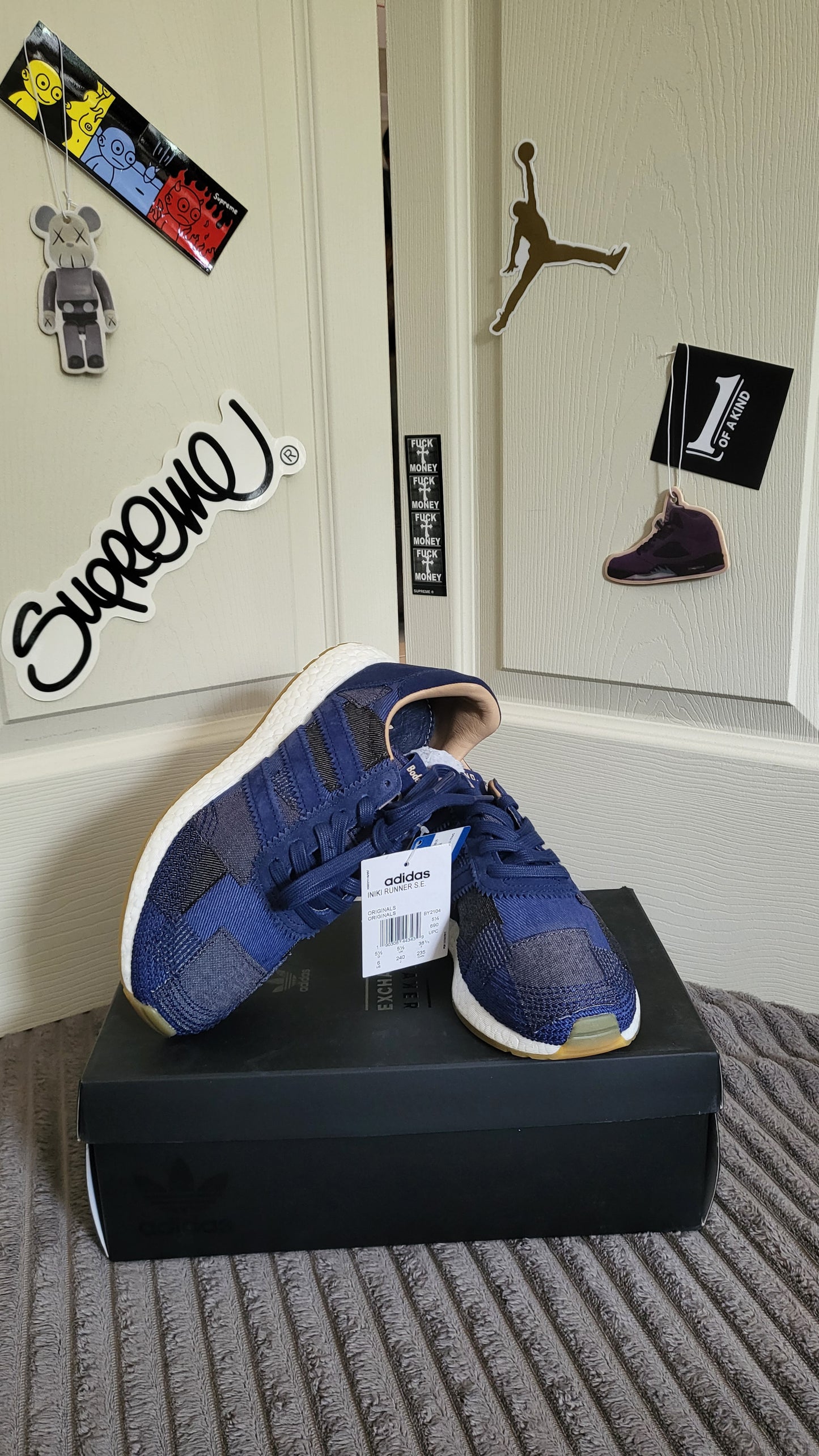 Front-facing shot of Adidas x Bedwin Iniki Runner SE in indigo on black shoebox, with wall stickers including Supreme, Jordan, and 1 of a Kind in the background.