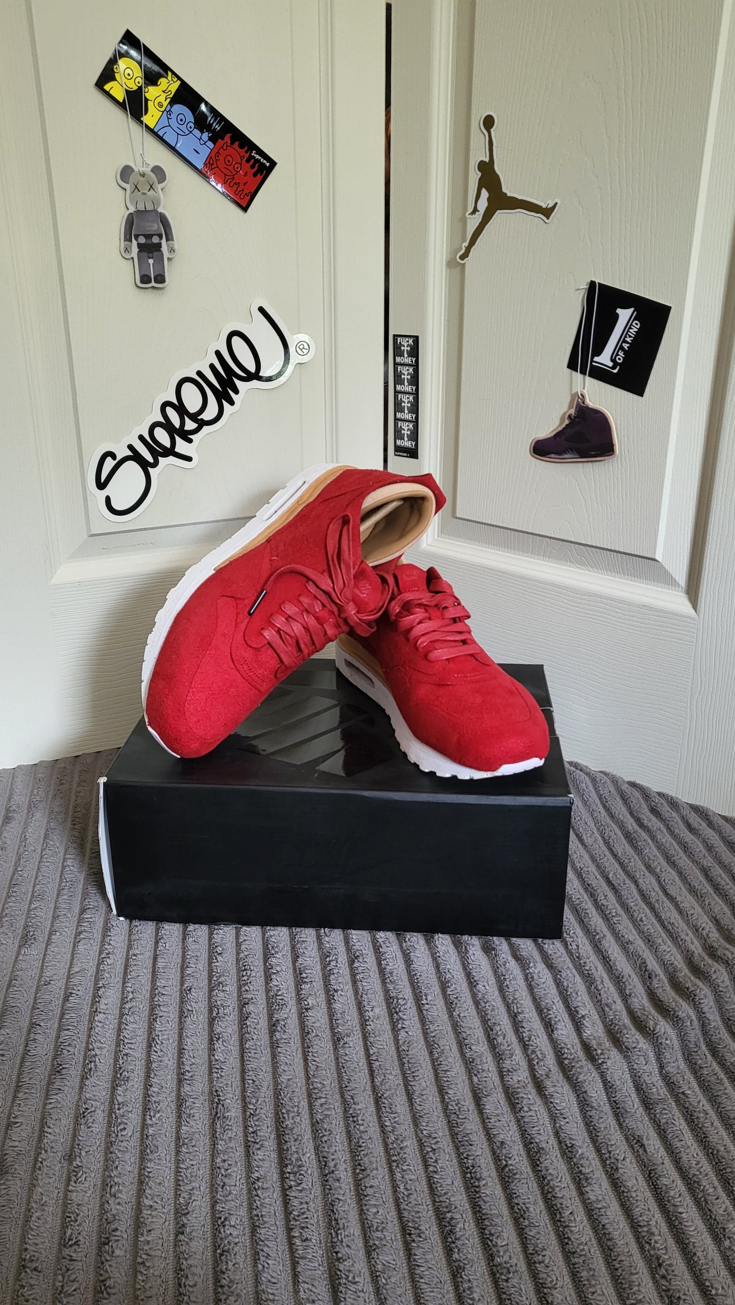 Nike Air Max 1 Royal in red suede on black box with streetwear stickers in background including Supreme and Jordan.