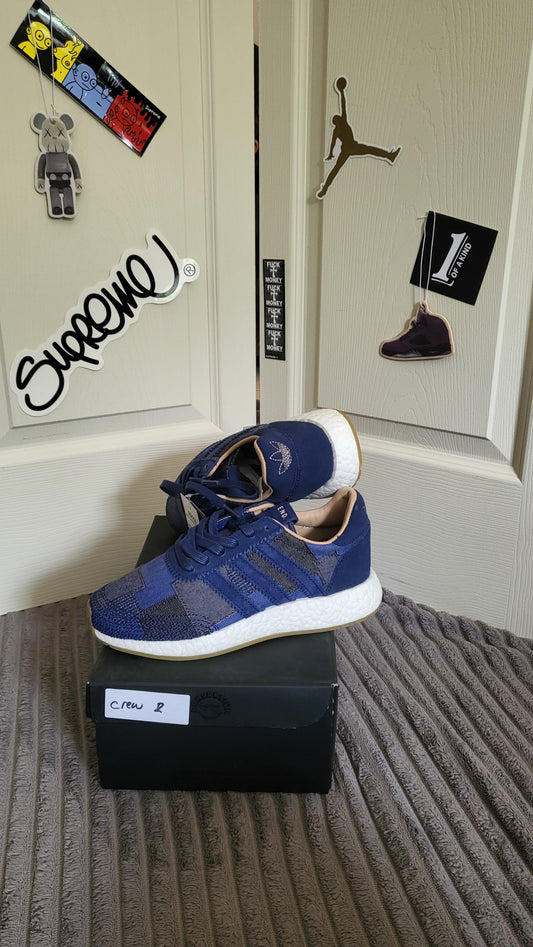 Adidas x Bedwin Iniki Runner SE in indigo displayed on a black shoebox with original tags, shown with streetwear stickers in the background.