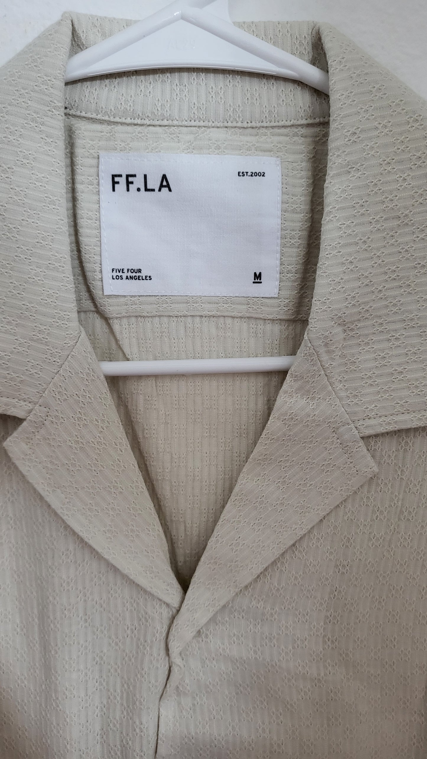 FF LA Cyrus cream shirt hanging against a white wall, photographed through a reflective surface with visible lighting flare and tag detail